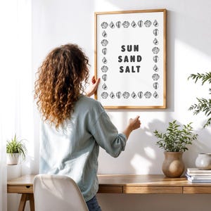 May include: A framed art print with the words "SUN SAND SALT" in bold black letters. The print is surrounded by a border of seashell illustrations. The frame is made of light brown wood. A person is hanging the framed art on a white wall.