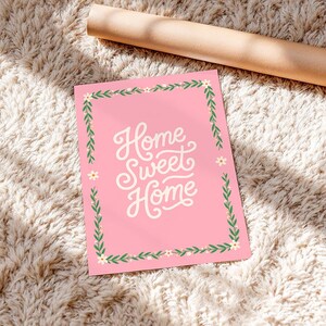 Könnte beinhalten: Eine rosa Karte mit den Worten "Home Sweet Home" in weißer Schrift. Die Karte ist mit einem grün-weißen Blumenrand umrahmt. Im Hintergrund liegt eine aufgerollte braune Papierrolle auf einer flauschigen, cremefarbenen Oberfläche.