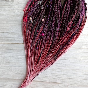 May include: A set of synthetic hair extensions featuring a gradient of deep burgundy, dark purple, and bright red tones. The hair is styled into numerous thin twists and braids, accented with small metal charms, pink beads, and thin decorative threads.