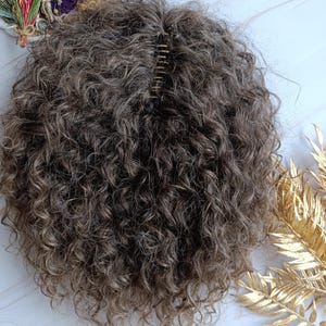 May include: A curly, brown hairpiece with a clip. The hairpiece has a natural texture and a mix of brown and blonde highlights. The clip is metal and designed to secure the hairpiece. This hair accessory is a wig or hair extension.