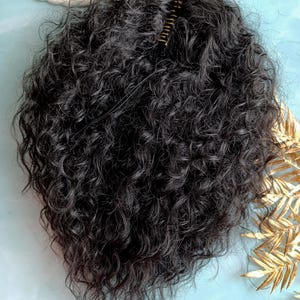 Può includere: Un'extension per capelli ricci scuri con attacco a pettine. I capelli sono di un nero intenso e sembrano sintetici. Il pettine è dorato e serve a fissare l'extension. L'extension è progettata per aggiungere volume e lunghezza ai capelli naturali.