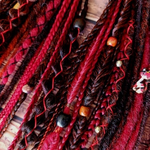 May include: Close-up of red and brown dreadlocks with various textures and embellishments. Braids, twists, and loose strands are interwoven, accented with wooden beads and red thread. The hair extensions have a natural, textured appearance.