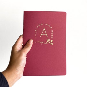 a women hand holding a simple notebook with blood red cover and a logo printed in gold foil