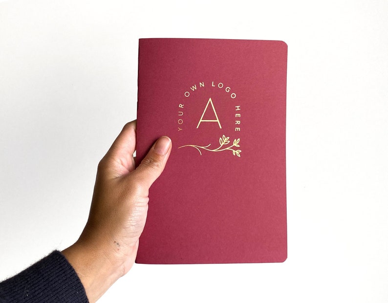 a women hand holding a simple notebook with blood red cover and a logo printed in gold foil