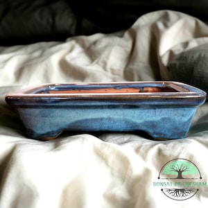 May include: A rectangular bonsai pot with a blue glaze and a brown rim. The pot has a low profile and sits on three small feet. The interior is a natural terracotta color. The pot is designed for bonsai trees.