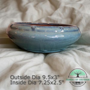 May include: A round, glazed ceramic bonsai pot with a light blue exterior and a reddish-brown interior. The pot measures 9.5 inches in diameter and 3 inches high on the outside, and 7.25 inches in diameter and 2.5 inches high on the inside. The pot is sitting on a white surface.