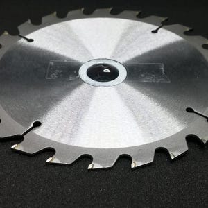 Può includere: Lama per sega circolare con finitura argentata e denti affilati. La lama ha un foro centrale e diversi fori più piccoli vicino al bordo esterno. La lama è progettata per tagliare legno o altri materiali.