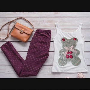 May include: A flat lay of a white vest top with a sequined teddy bear motif, burgundy polka dot trousers, a brown crossbody bag, and pink trainers. The items are arranged on a light wood surface.