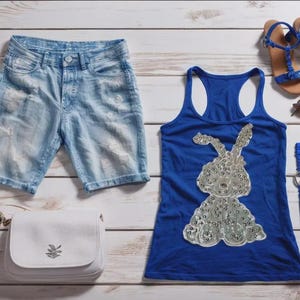 Könnte beinhalten: Sommerliche Kleidung und Accessoires, draufsicht. Hellblaue Jeansshorts, ein königsblaues Tanktop mit silbernem Pailletten-Hasenmuster, blaue Sandalen, eine weiße Handtasche, Sonnenbrille, Uhr und Armbänder.