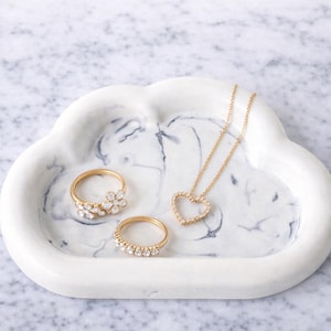 May include: A white cloud-shaped tray with a marble design holds gold rings and a necklace. The rings feature floral and gemstone designs. The necklace has a heart pendant with gemstones.