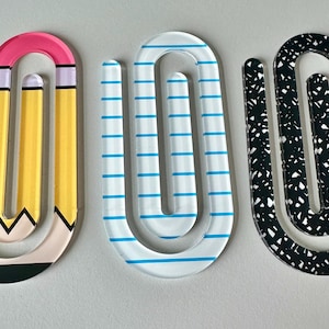 May include: Three large paperclip-shaped bookmarks made of clear acrylic. The first features a yellow pencil design with a pink eraser and black tip. The second is clear with horizontal blue lines. The third has a black and white speckled pattern.
