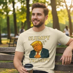 Könnte beinhalten: Beiges T-Shirt mit dem Text "SLUMPY TRUMPY" und einem Cartoon-Bild eines schlafenden Mannes mit blonden Haaren. Das T-Shirt wird von einer Person getragen, die auf einer Holzbank in einem Park sitzt.