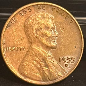 Puede incluir: Una moneda de centavo Lincoln de 1955 D de color cobre. El anverso presenta un perfil de Abraham Lincoln, con las inscripciones "IN GOD WE TRUST" y "LIBERTY". El año "1955" es visible, junto con la marca de ceca "D".