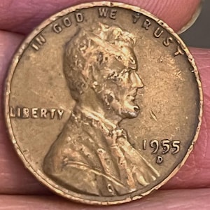 May include: A close-up of a copper-colored 1955 D Lincoln penny. The coin features the profile of Abraham Lincoln, the word "LIBERTY", and the inscription "IN GOD WE TRUST". The year "1955" is visible.