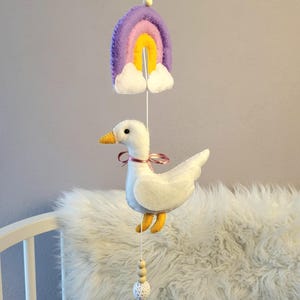Könnte beinhalten: Handgefertigtes Baby-Mobile mit einer weißen Filz-Gans, gelbem Schnabel und Füßen sowie einem rosa Band. Über der Gans befindet sich ein Regenbogen aus Filz in Lila, Rosa und Gelb. Das Mobile hängt an einer Schnur mit Holzperlen und einem Kristall.