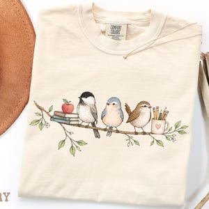May include: An ivory t-shirt featuring a graphic of three birds perched on a leafy branch. The illustration includes a stack of books with a red apple, a black-capped bird, a blue bird, a brown bird, and a mug filled with pencils and pens.