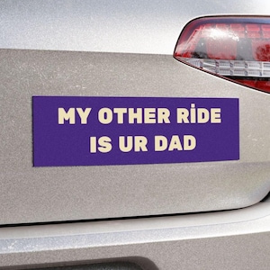 May include: A purple bumper sticker with the text "MY OTHER RIDE IS UR DAD" in white letters is affixed to the back of a silver car. The car's taillight is visible on the right side of the image.