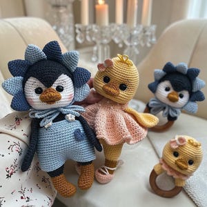 May include: Crocheted penguin and duckling dolls in blue, black, yellow, and pink. The penguin wears blue overalls and a flower-shaped hat. The duckling wears a pink dress. A duckling rattle is also visible.