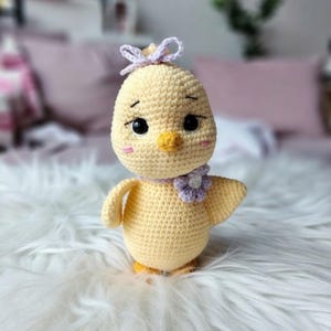 Może przedstawiać: Ręcznie robione, żółte szydełkowe pisklę z lawendową kokardą na głowie i lawendowym kołnierzem z kwiatów. Pisklę ma czarne oczy, żółty dziób i różowe policzki. Pisklę stoi na białej, puszystej powierzchni.