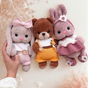 Puede incluir: Tres animales de peluche de ganchillo hechos a mano: dos conejos y un oso. Los conejos son de color rosa claro con lazos y vestidos. El oso es marrón con peto amarillo. Todos tienen ojos negros y están sobre un fondo claro.