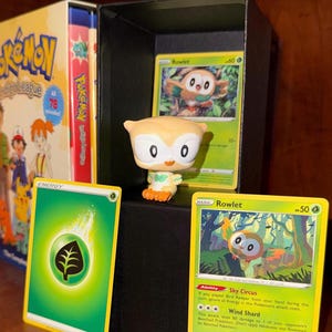 May include: A collection of Pokémon merchandise featuring a Rowlet figure and trading cards. The cards include an Energy card and two Rowlet cards with forest backgrounds. A Pokémon Indigo League box set is visible in the background.