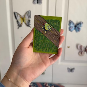 May include: A small, rectangular artwork featuring a colorful insect on a brown log against a green background. The artwork is held in a hand, with butterfly decorations on the white door behind it. The insect has a yellow body, red head, and blue and yellow legs.