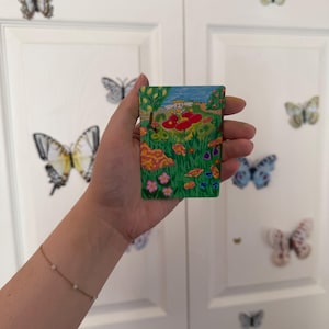 May include: A rectangular card featuring a colorful illustration of a white cartoon character with long ears sitting on a large red flower in a green meadow filled with various small flowers, trees, and a blue sky with a yellow sun in the background.