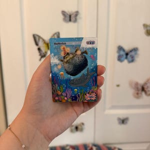 May include: A colorful card featuring an underwater scene with a pufferfish, a shark, and coral. The card is labeled "Blufferfish" and includes other text and symbols. The card is held in a hand, with butterflies on the background.