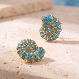 Op de afbeelding: Een paar goudkleurige oorbellen in de vorm van nautilusschelpen, met een textuur die dicht bezet is met kleine, helderblauwe turkooiskleurige steentjes en een grotere, gladde turkoois cabochon in het midden van elke spiraal.