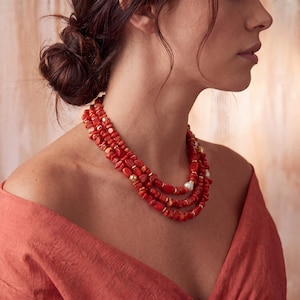 May include: A multi-strand necklace featuring polished red coral chips and smooth, round gold-tone beads. The necklace is layered with three distinct strands, incorporating small white pearls interspersed throughout the vibrant red coral segments.