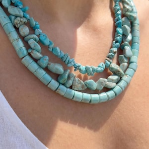 Può includere: Una collana a più fili con pietre color turchese di varie forme e dimensioni. Il design include perline cilindriche lisce, pepite irregolari levigate e pezzi più piccoli simili a scaglie, tutti infilati insieme in una disposizione a strati.