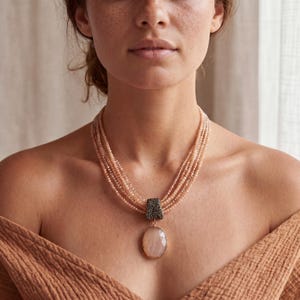 May include: A multi-strand necklace featuring pale peach-coloured faceted beads arranged in several layers. The strands are gathered by a dark, textured rectangular clasp, from which hangs a large, smooth, oval-shaped pale peach gemstone pendant.
