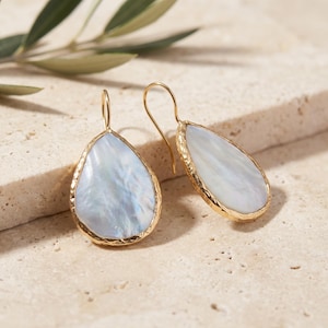 Peut inclure: Une paire de boucles d'oreilles en forme de goutte avec des centres en nacre blanche irisée encastrés dans des bords en métal doré texturé. Chaque boucle d'oreille est suspendue à un crochet doré simple, révélant une surface lisse et réfléchissante.
