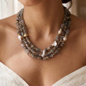 Op de afbeelding: Een meerstrengs ketting met grijze labradoriet chipkralen, afgewisseld met kleine goudkleurige metalen tussenstukjes en diverse onregelmatige witte zoetwaterparels. De ketting bestaat uit drie afzonderlijke strengen die om de hals vallen.