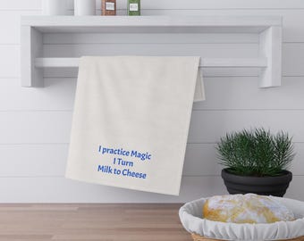 May include: A white kitchen towel with blue text that reads "I practice Magic I Turn Milk to Cheese" hangs from a white shelf. The shelf holds jars and spices. A basket of bread and a potted plant are on the counter.