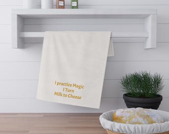 May include: Off-white kitchen towel with the text "I practice Magic I Turn Milk to Cheese" in gold lettering. The towel hangs on a white shelf with jars and spices. A loaf of bread sits in a wicker basket on a wooden countertop.