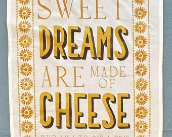 May include: A white rectangular tea towel featuring a mustard yellow floral border and the text SWEET DREAMS ARE MADE OF CHEESE WHO AM I TO DIS A BRIE printed in bold mustard yellow and dark brown lettering centered on the fabric surface.