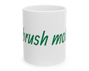 May include: A white ceramic mug with the words "brush mo" printed in green cursive script. The mug is cylindrical and appears to be empty. The mug is a standard size for coffee or tea.