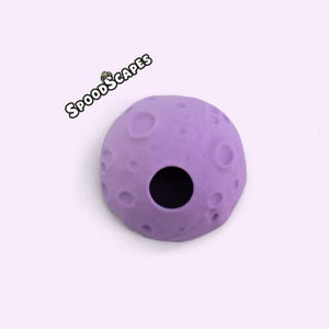 Puede incluir: Un objeto esférico de color morado claro con una superficie texturizada que presenta múltiples cráteres circulares y una única abertura circular grande y oscura. El texto SPOODSCAPES está impreso en letras blancas y negras sobre el objeto.