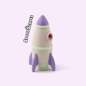 Könnte beinhalten: Eine 3D-gedruckte Raketenfigur in Creme, Lila und Hellgrün. Die Rakete hat eine lila Nasenspitze, ein rosa umrandetes Bullauge mit einer kleinen Spinne darin und drei lila Flossen an der Basis. Der Text SPOODSCAPES ist an der Seite aufgedruckt.