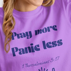 Puede incluir: Una camiseta de color lila con el texto en azul marino "Pray more Panic less". La camiseta también presenta el texto "1 Thessalonians 5:17" y un gráfico de manos sosteniendo un sol.
