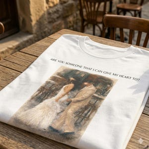 Op de afbeelding: Een wit t-shirt met een bedrukte afbeelding van een persoon in een witte jurk en een persoon in een beige pak die samen staan. Boven de afbeelding staat de tekst: ARE YOU SOMEONE THAT I CAN GIVE MY HEART TO? Het t-shirt heeft een ronde hals.