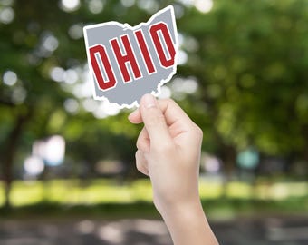 May include: A sticker in the shape of the state of Ohio, with the word "OHIO" in red letters outlined in white. The sticker is held up against a blurred green background.