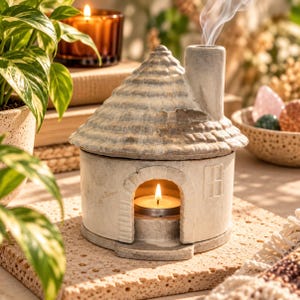 May include: A ceramic house-shaped oil burner with a textured off-white finish, featuring a shingled roof, a chimney emitting smoke, and an arched front opening containing a lit tea light candle to warm essential oils.
