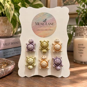 May include: A set of six small turtle-shaped pins attached to a white display card featuring the text Muse Lane Collective, earthy rituals, subtle rebellion. The turtles are colored in shades of purple, green, and orange with a textured finish.