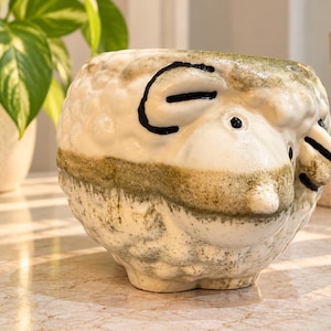 May include: A ceramic planter shaped like a sheep with a textured, off-white surface featuring green-toned weathering and hand-painted black details for the eyes and ears. The vessel has a rounded body and a wide opening at the top.