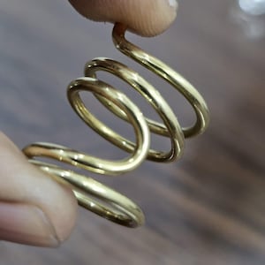 Può includere: Un anello a forma di spirale color oro. L'anello è tenuto tra due dita e ha una superficie liscia e lucida. Il design presenta più anelli, creando un aspetto unico. L'anello sembra essere fatto di metallo.