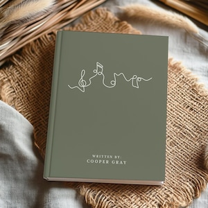 May include: A sage green journal featuring a white line drawing of musical notes. The cover also displays the text "WRITTEN BY: COOPER GRAY". The journal is placed on a hessian surface.