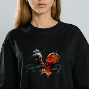 May include: Black t-shirt featuring a graphic design of two skulls with a butterfly and plant elements. The artwork has a vibrant color palette with red, orange, and blue accents. The shirt has short sleeves and a relaxed fit.