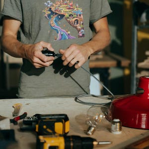 May include: A person wearing a gray t-shirt with a colorful fantasy landscape graphic. The image shows a workshop setting with tools, a red lamp, and a lightbulb. The person is working on a black object with a silver cable.